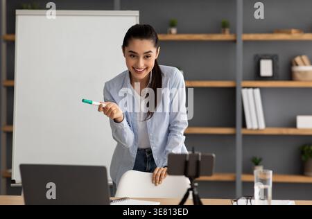 Arab female tutor holding marker explaining lesson at whiteboard teaching online using smartphone while standing near table with laptop at home Stock Photo