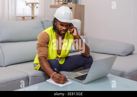 Black male engineer with twists wearing hard hat and safety vest having phone call, using laptop, and taking notes while sitting on sofa at home of... Stock Photo
