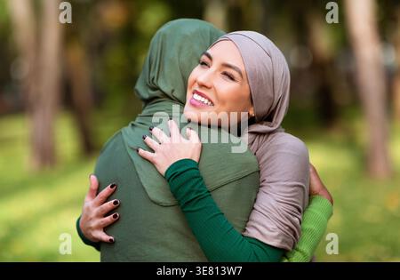 Smiling Middle Eastern and Black women wearing hijabs embracing each other while standing in city park during daytime Stock Photo