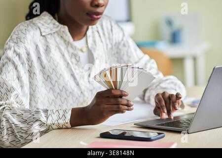 Black female designer with braids holding color palette working on laptop while sitting at table in office Stock Photo