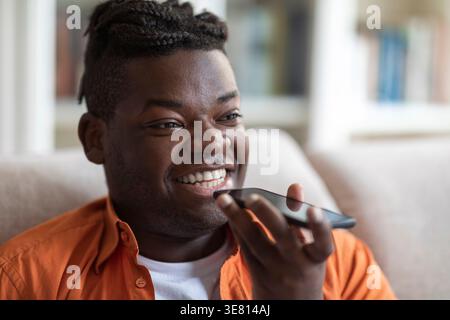 Smiling Black man with braids recording voice messages sitting on sofa in living room at home Stock Photo