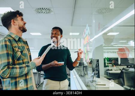 Black businessman writing his ideas on sticky notes and discussing project plan with caucasian male colleague holding tablet laughing while standin... Stock Photo