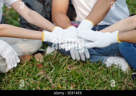 Group of Asian volunteers stacking hands with gloves sitting on grass in park during daytime Stock Photo