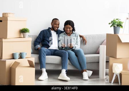 Black husband and wife using tablet together while sitting on sofa in living room near unpacked boxes at home Stock Photo