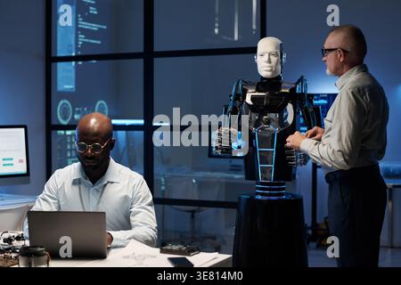Black male computer engineer with eyeglasses working on laptop while sitting at table in front of his colleague and robot in laboratory Stock Photo