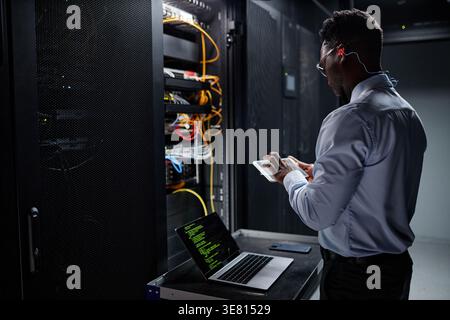 Black male network engineer using tablet and laptop while standing and setting up servers in data center Stock Photo