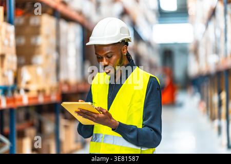 Black male supervisor with dreads wearing safety vest and hard hat doing inventory on tablet while standing in logistic warehouse Stock Photo