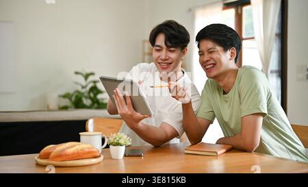 Cheerful Asian male friends watching funny videos on tablet while sitting at table and studying together at home Stock Photo