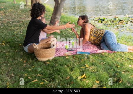 Hispanic middle-aged mother blowing bubbles with her daughters while lying on grass by the lake during daytime Stock Photo