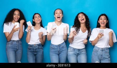 Cheerful diverse group of women wearing white shirts and jeans using smartphones standing against blue background in studio Stock Photo