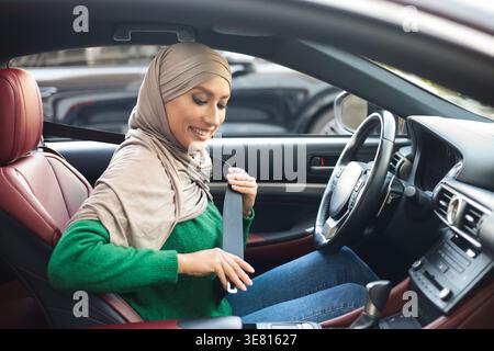 Smiling Middle Eastern woman wearing hijab putting on seatbelt while sitting inside car during daytime Stock Photo
