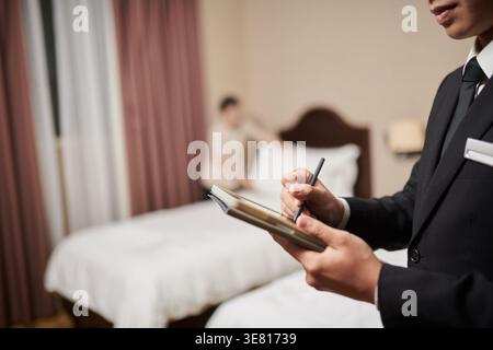 Cropped image of Asian male manager taking notes standing near female assistant making bed in hotel room Stock Photo