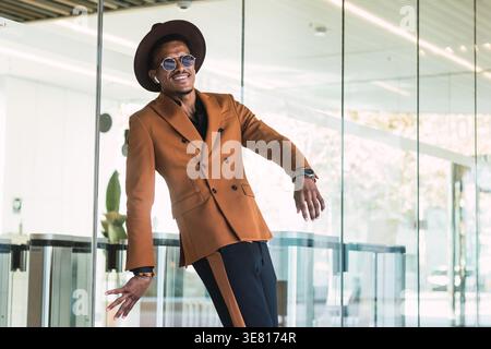 Hispanic man wearing brown suit, sunglasses, and fedora hat standing and leaning on glass wall outside the building in city during daytime Stock Photo