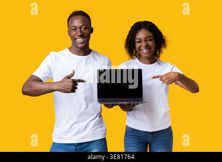 Smiling Black couple pointing fingers at laptop while standing against yellow background in studio Stock Photo