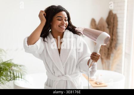 Black woman in bathrobe blow drying hair standing in bathroom at home Stock Photo