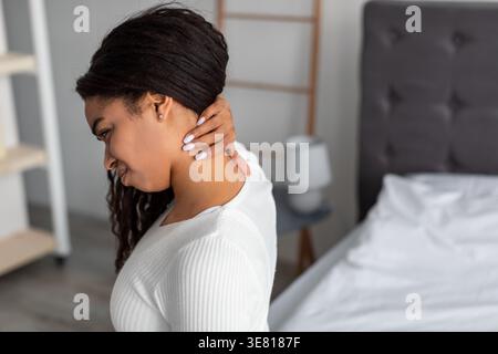 Black woman massaging her neck sitting o bed in bedroom at home Stock Photo