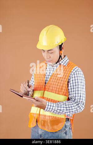Asian male civil engineer in vest and hardhat writing on tablet standing against brown background in studio Stock Photo