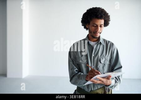 Black male contractor in gray uniform taking notes while standing and inspecting construction site Stock Photo
