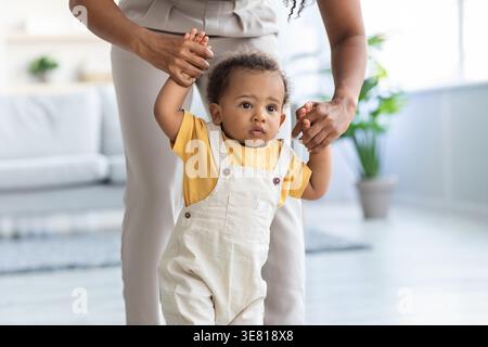 Black baby boy wearing jumpsuit walking with mother holding his hands in living room at home Stock Photo