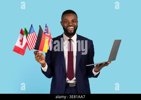 Smiling Black businessman in suit holding laptop and different flags while standing against blue background in studio Stock Photo