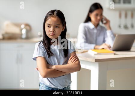 Sad Asian girl with crossed arms looking away standing near her mother having phone call working on laptop while sitting at table in kitchen at home Stock Photo