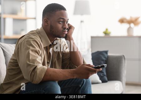 Bored Black man with face leaning on hand holding remote control watching TV while sitting on sofa in living room at home Stock Photo