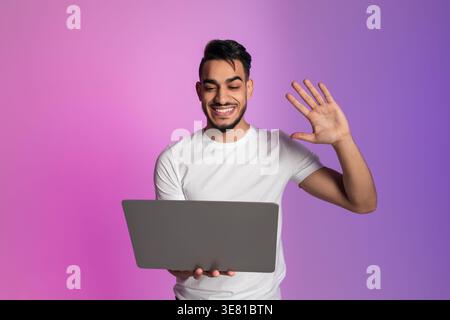 Arab man with beard having video call on laptop and waving hand standing against pink and purple gradient background in studio Stock Photo