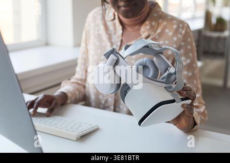 Black businesswoman holding virtual reality headset working on computer while sitting at desk in office Stock Photo