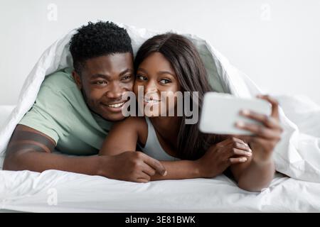 Black couple under bed sheet taking selfie using smartphone while lying down on bed at home Stock Photo