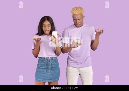 Black couple using smartphones showing hand gestures while standing together against purple background in studio Stock Photo