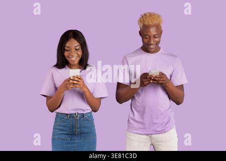 Black couple using smartphones while standing against purple background in studio Stock Photo