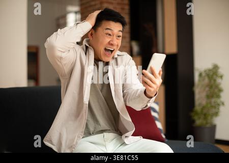 Shocked Asian man holding smartphone and touching head sitting on sofa in living room at home Stock Photo