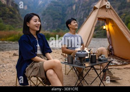 Smiling Chinese couple looking away sitting on folding chairs having breakfast near river and mountains during camping Stock Photo