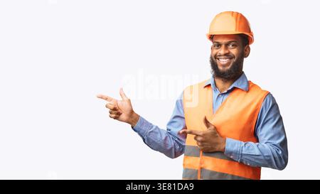 Black male worker wearing safety helmet pointing fingers sideways while standing against white background in studio Stock Photo