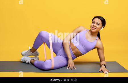 Black woman in sportswear doing exercise with resistance band while lying on yoga mat against yellow background in studio Stock Photo