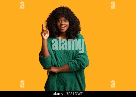 Black woman with curly afro pointing finger up while standing against yellow background in studio Stock Photo