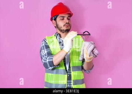 Arab male construction worker wearing safety vest and red hardhat cleaning his goggles with handkerchief while standing against pink background in ... Stock Photo