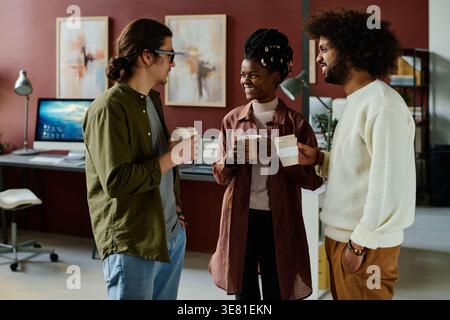 Black female office worker with braids having coffee with mixed-race and caucasian male colleagues discussing project details while standing close ... Stock Photo