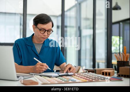 Asian middle-aged male interior designer using digital tablet sitting at the table with materials in office Stock Photo
