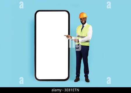 Cheerful Black male engineer wearing hard hat and safety vest pointing fingers at big smartphone standing against blue background in studio Stock Photo