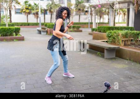 Smiling Hispanic woman with curly hair wearing black top and blue jeans dancing in city park during daytime Stock Photo