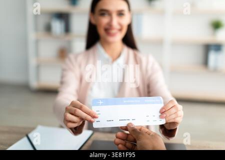 Caucasian female travel agent giving ticket to client while sitting at table in tour agency office Stock Photo