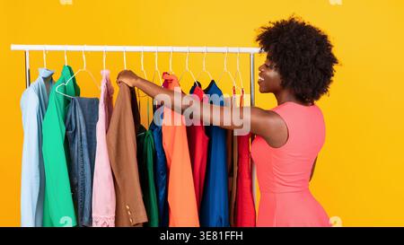Black woman choosing clothes from rack while standing against yellow background in studio Stock Photo