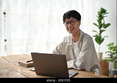 Smiling Asian male in eyeglasses sitting at table with laptop  at home Stock Photo