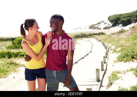 Smiling Black man and caucasian girlfriend embracing and looking at each other while standing on boardwalk near beach on a summer day Stock Photo