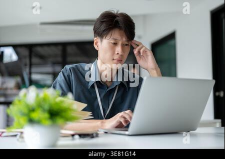 Confused Asian male office worker working on laptop sitting at desk in office Stock Photo