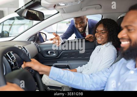 Black salesman in business suit bending forward on car window and talking with couple sitting inside the car in showroom Stock Photo