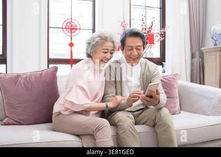Smiling senior Chinese couple using smartphone together while sitting on sofa at home Stock Photo