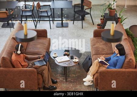 High-angle view of Mixed-race woman writing on pad while Black man with headphones working on his laptop sitting on couch in cafe Stock Photo