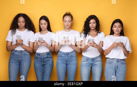 Diverse group of women wearing white shirts and jeans using smartphones standing against yellow background in studio Stock Photo